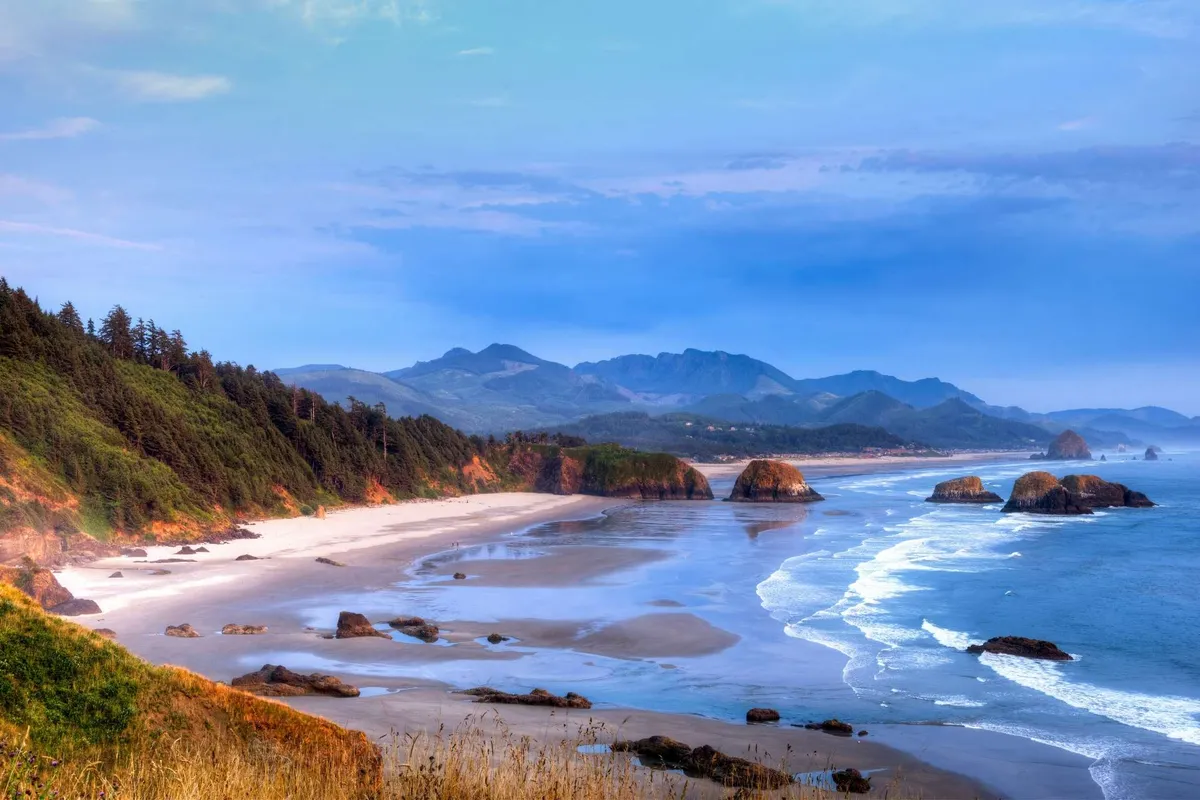 A stunning view of the Oregon Coast featuring a sandy beach, rugged rock formations, the ocean, and distant mountains framed by trees. The tranquil setting invites outdoor exploration such as beachcombing, hiking, and photography.