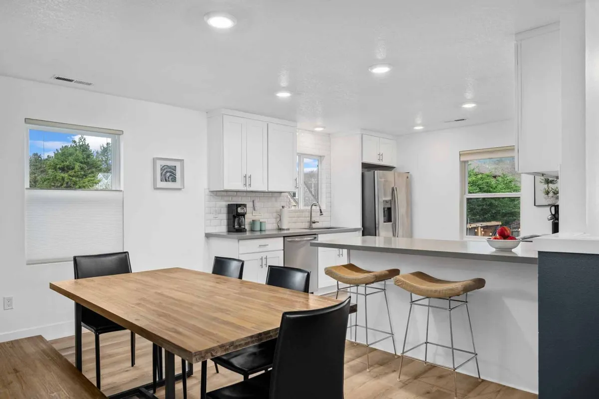 A welcoming dining area showcasing a large dining table, bar stools at the kitchen counter, and ample natural light from surrounding windows. The setup is ideal for group meals, casual brunches, or evening celebrations, offering both comfort and functionality.