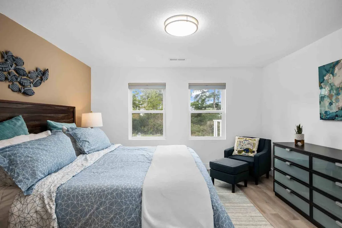 A bright and airy bedroom with patterned bedding, two large windows filling the room with natural light, an accent chair, and decorative wall art. The space appears relaxing and inviting, with a cozy reading nook by the window.