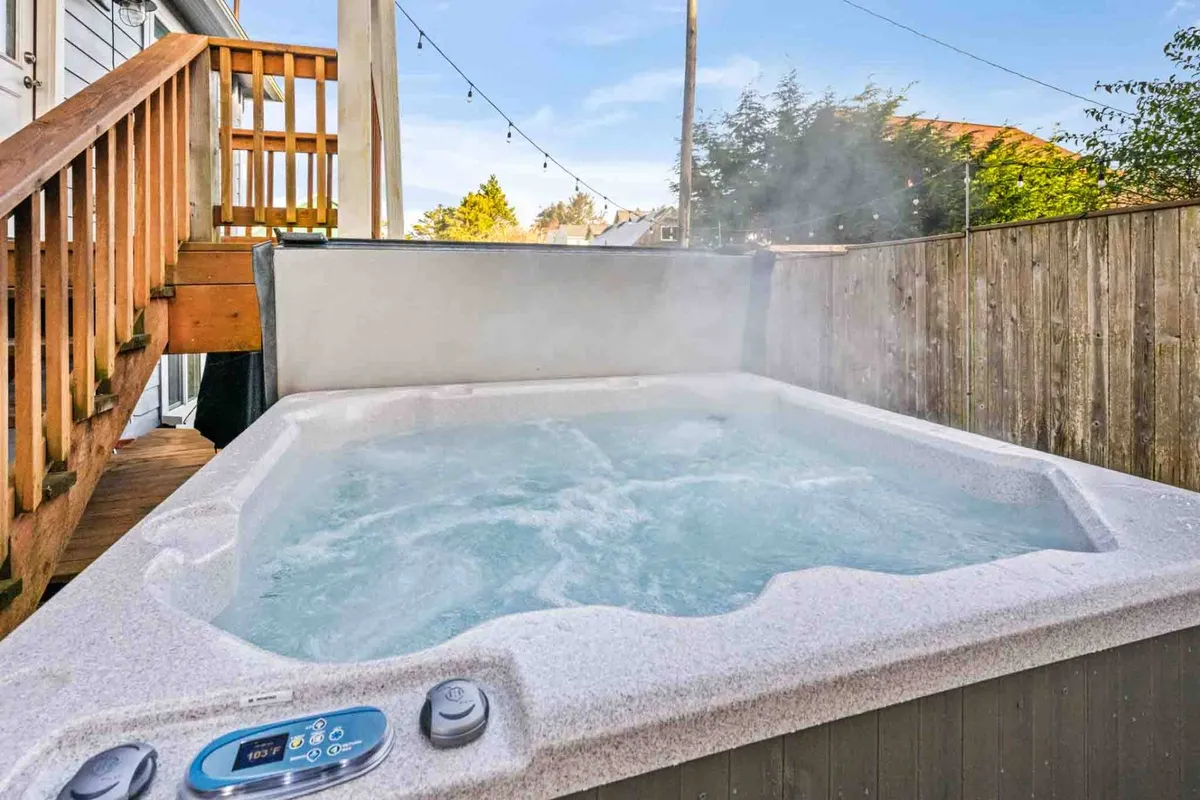 A serene hot tub sits on a wooden deck, surrounded by lush greenery. The area is designed for relaxation, with ample space for unwinding in privacy while still enjoying the outdoors. The lighting suggests it’s a perfect spot for both sunset viewing and stargazing at night.