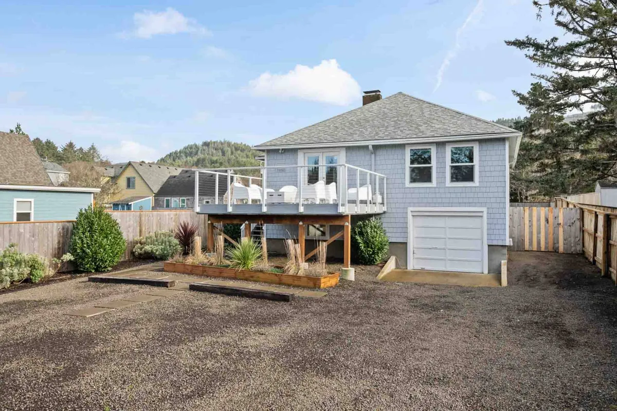 A spacious deck with comfortable seating overlooks a beautifully landscaped garden area. The deck offers views toward the surrounding natural scenery and provides a welcoming spot for outdoor relaxation, morning coffee, or evening gatherings. The garage entrance is visible, giving a sense of the property's layout and ease of access.