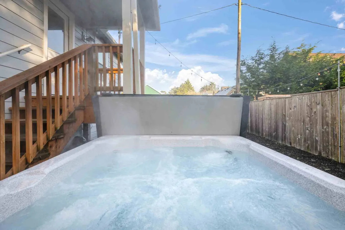 This image shows a cozy outdoor hot tub area surrounded by a wooden fence, with inviting string lights overhead and wooden stairs leading down. The scene is tranquil and inviting, perfect for unwinding after a day at the beach or exploring downtown. The mood is serene, suggesting an ideal spot for group relaxation under the stars.
