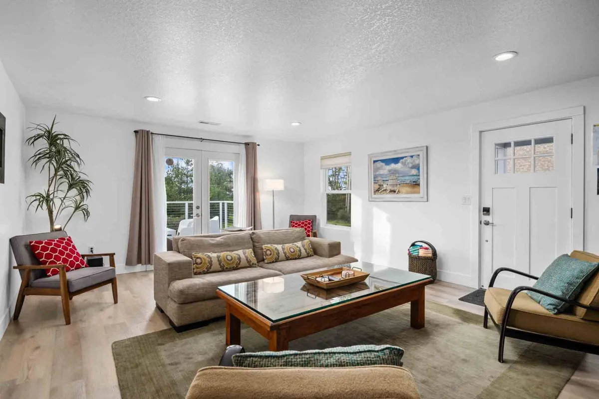 A bright and cheerful living space featuring a sofa with colorful cushions, glass coffee table, large windows letting in abundant natural light, vibrant artwork, and an indoor plant. The space is arranged for relaxation and socializing.