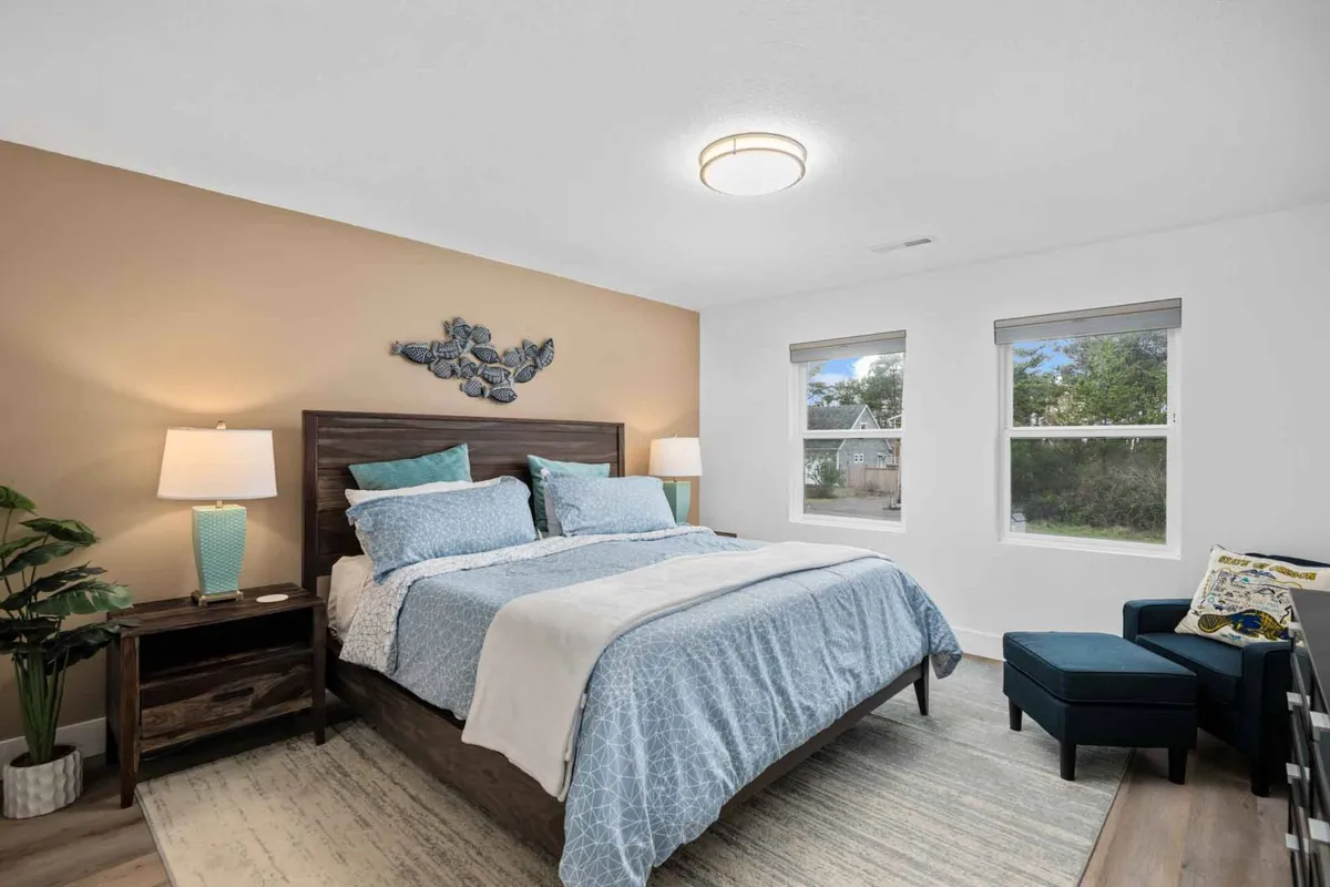 A serene bedroom featuring a bed with blue bedding, decorative wall art, a nightstand with a lamp, a potted plant, and an armchair beside a window. Sunlight streams in, creating a calm and peaceful atmosphere—perfect for relaxing or enjoying a quiet morning coffee.